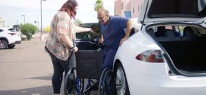 A senior citizen in a wheelchair is being transported into the trunk of a white car.