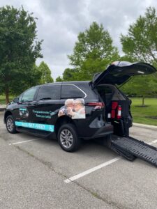 A black van with "Compassionate Care Transportation" written on the side is parked in a lot with the rear door open and a ramp extended, adjacent to a grassy area with trees. This wheelchair transportation company ensures accessibility and ease for all its passengers.