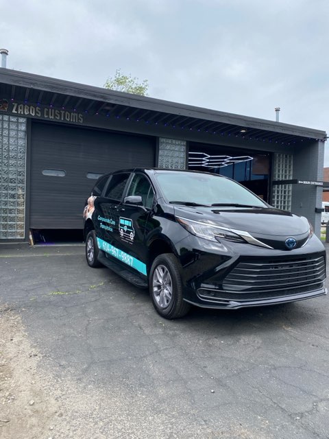 A black van with business graphics offering Wheelchair Transportation Services is parked in front of a building labeled "Zaggs Customs." The building features a glass block design at the entrance.