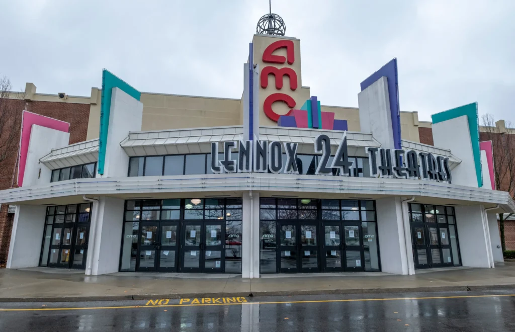 Front view of Lennox 24 AMC theater with a modern facade, colorful signage, and multiple entrances. Wet pavement suggests recent rain.