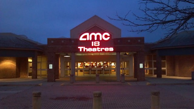 Front view of an AMC 18 Theatres building at dusk, with an illuminated sign and a paved entrance.