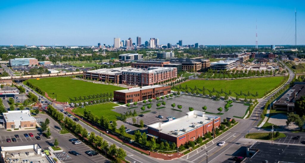 Aerial view of a city skyline with green fields, brick buildings, parking lots, and roads in the foreground, under a clear blue sky.