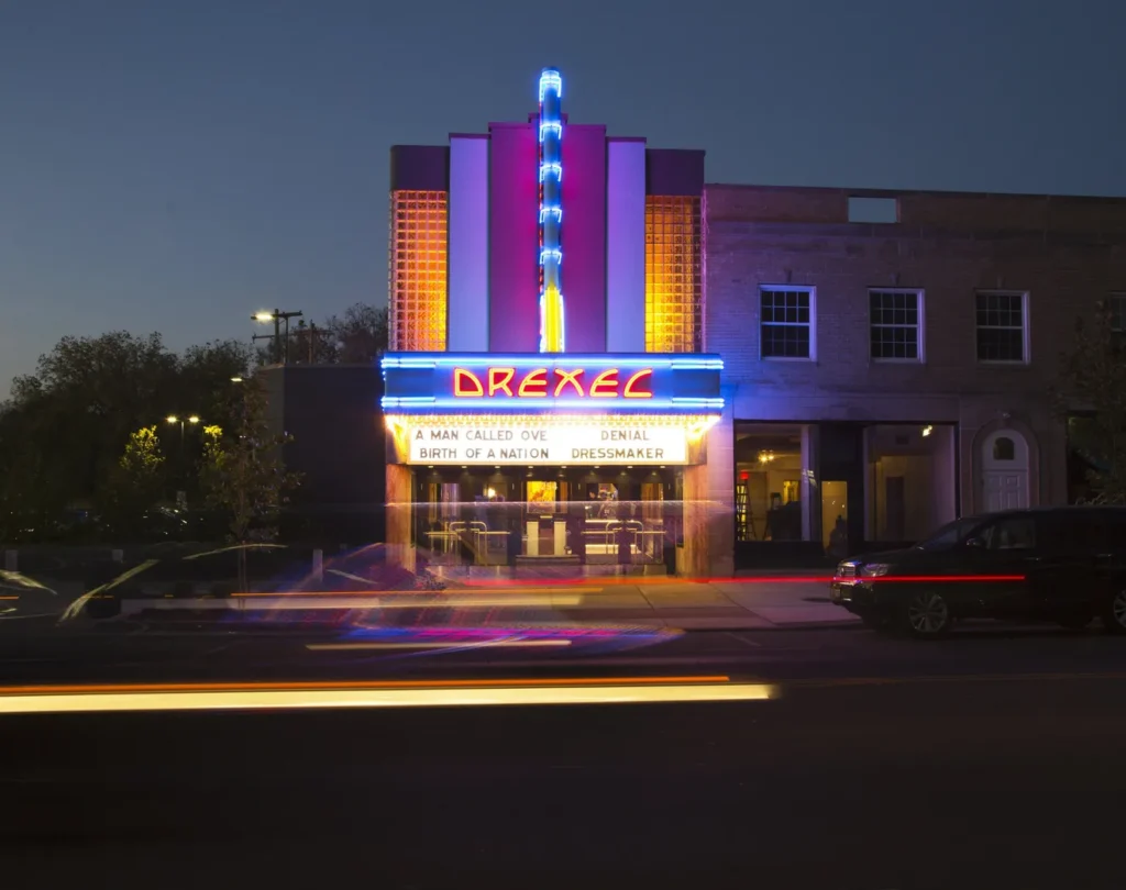 A vintage movie theater with neon lights displays films on its marquee. Cars pass by on the street at dusk.