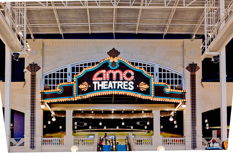 Entrance of an AMC Theatres, featuring a large illuminated sign above escalators, set in a brightly lit interior with high ceilings.