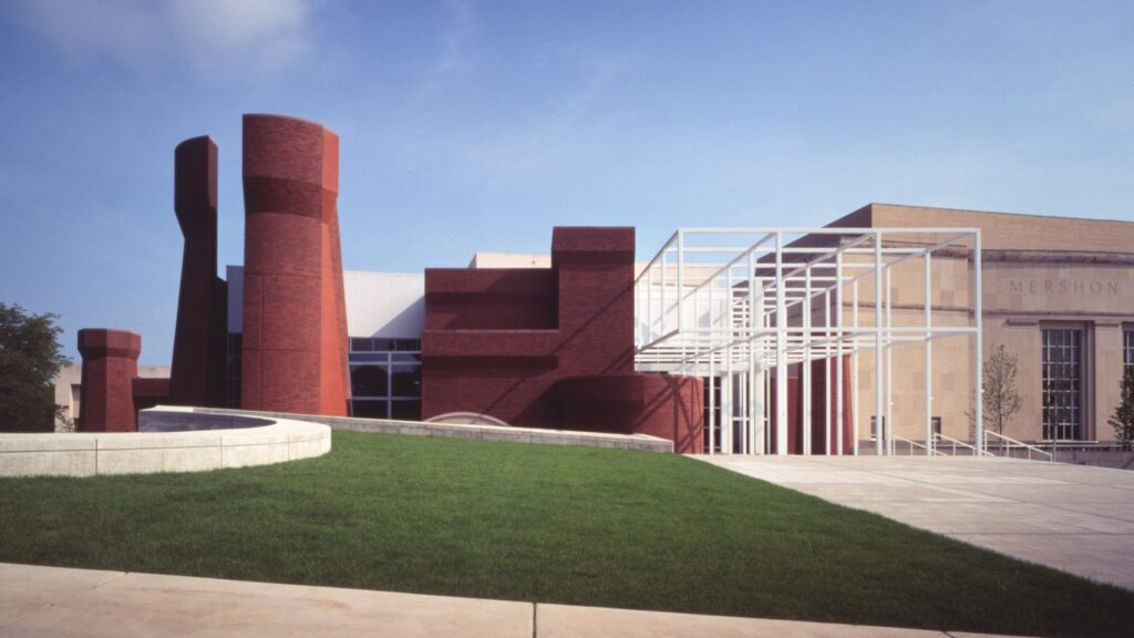 Modern building with red brick and geometric white structures, adjacent to Mershon auditorium entrance, surrounded by grass and pavement under a clear sky.