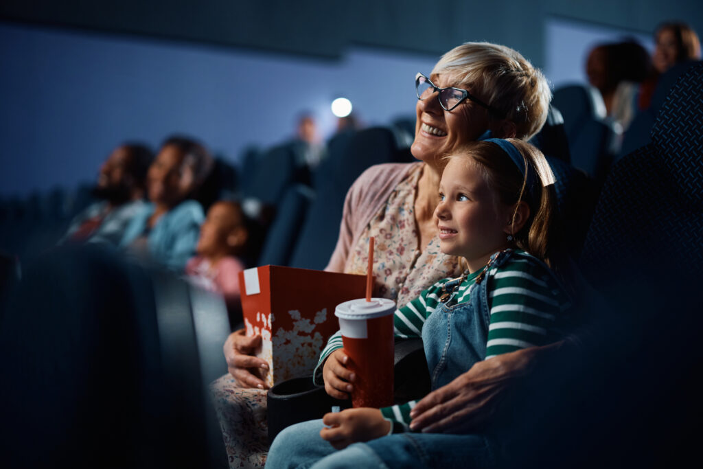 A woman and young girl sit together in a movie theater, smiling. The woman holds a popcorn box, and the girl has a drink. Other people are visible in the background.