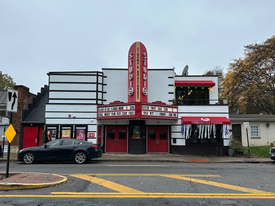 Black car parked in front of a vintage-style theater with a red marquee. To the right, a sign reads "Francesco's Pizzeria" with outdoor seating.