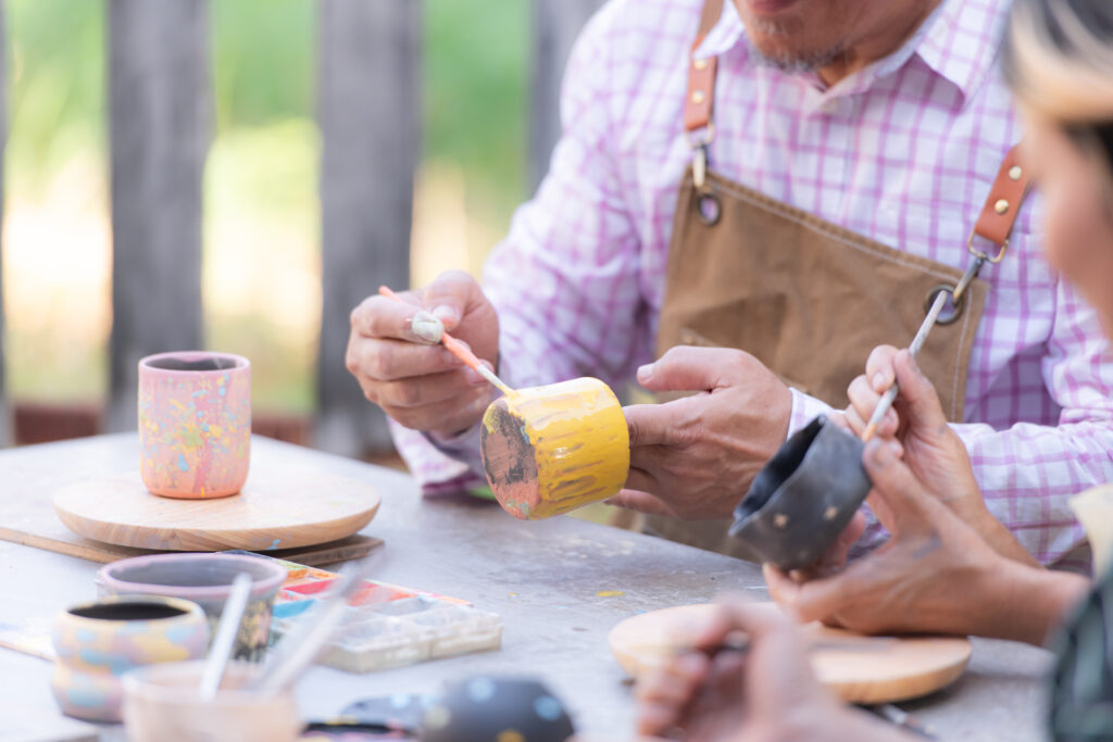 People painting ceramic mugs outdoors, using small brushes and various colors.