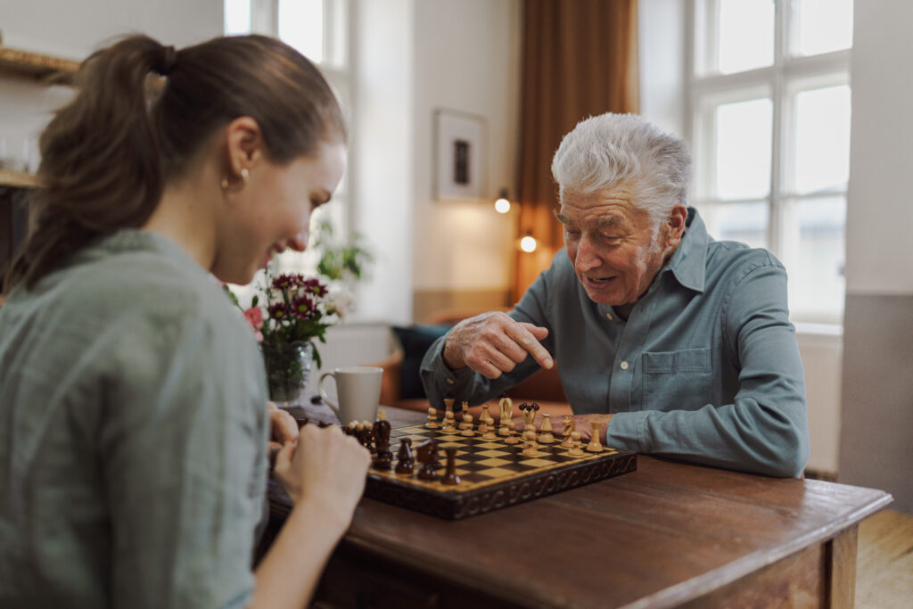 Older man and younger woman play chess at a wooden table in a sunlit room, with a vase of flowers and a coffee cup nearby.