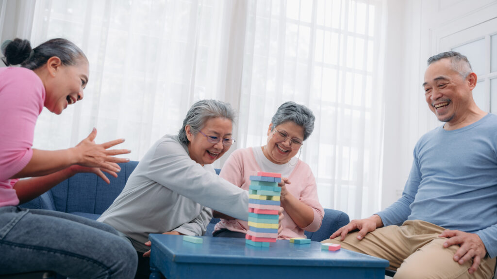 Four older adults play a game of block stacking on a blue table. They smile and seem engaged, with a bright window in the background.