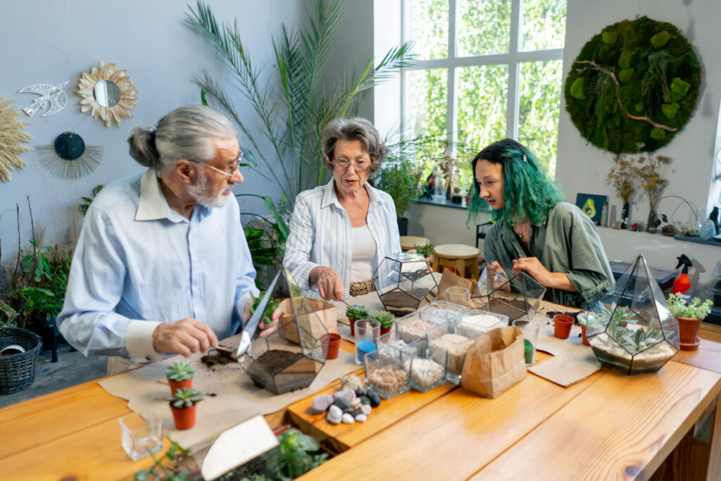 Three people creating terrariums at a table filled with plants and supplies, in a well-lit room with large windows and green decor.