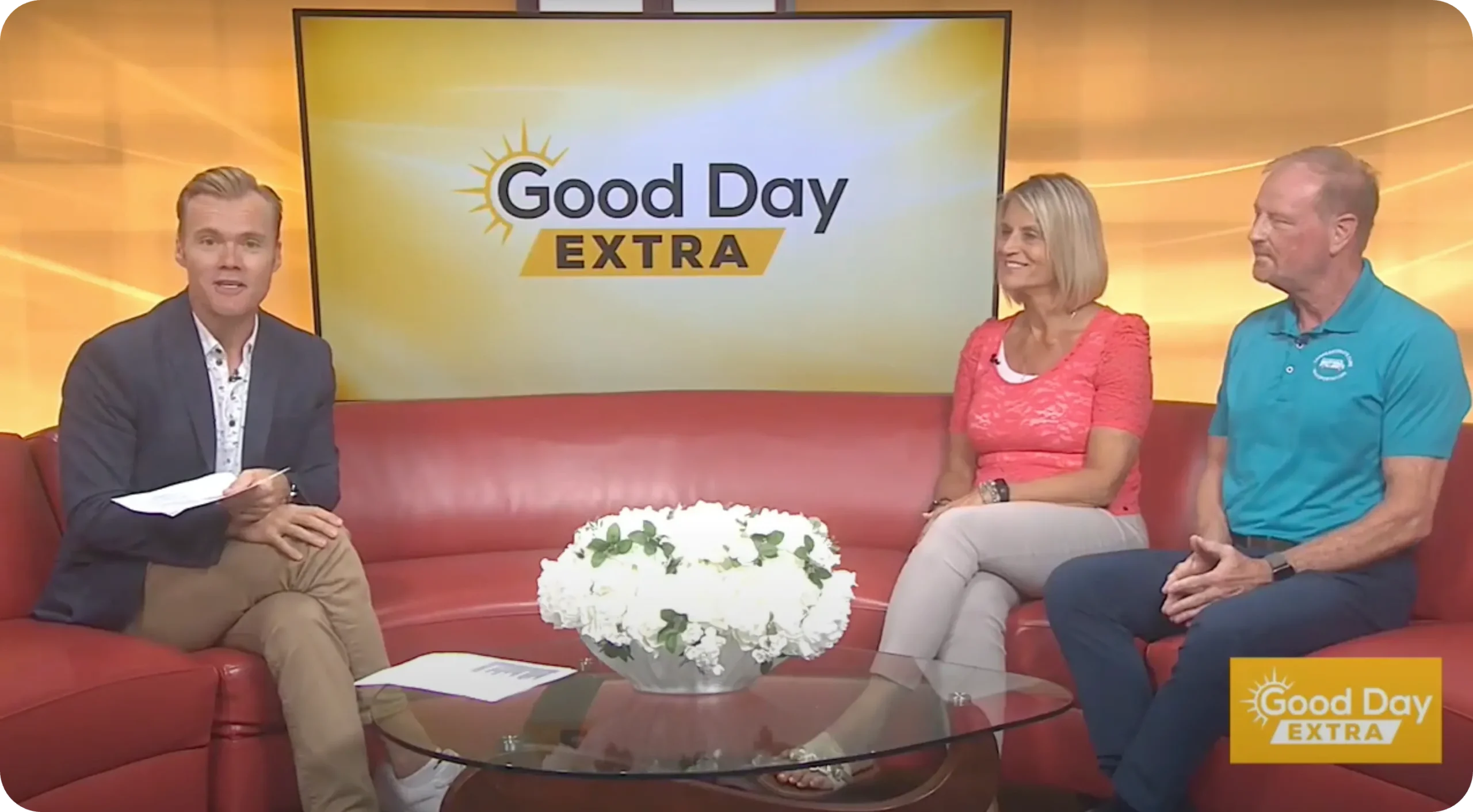 Three people sit on a red couch during a "Good Day Extra" TV segment, discussing local wheelchair transportation services, with a bouquet of white flowers on the glass coffee table in front of them.