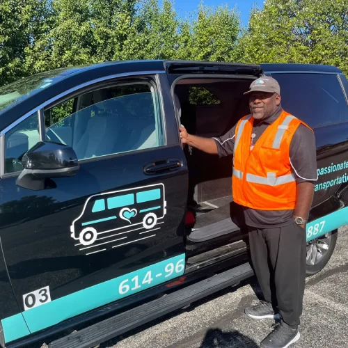 Man in orange safety vest stands beside a black van with a transportation company logo, holding the open door, in a sunny outdoor parking lot with trees in the background—ready to provide reliable wheelchair transportation services for those searching for “wheelchair transportation services near me.”.