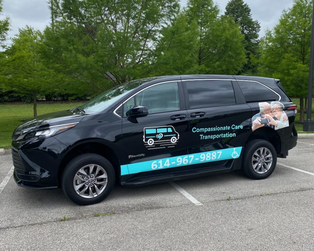 A black van with "Compassionate Care Transportation" branding and contact number 614-967-9887 is parked in a lot with trees and greenery in the background, offering reliable wheelchair transportation services near me.